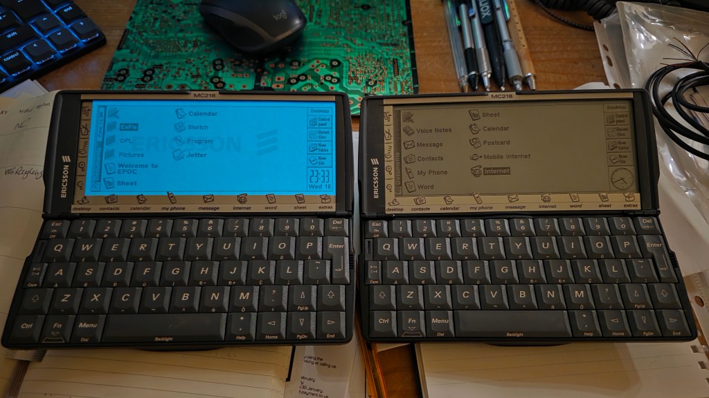 Two Ericsson MC218 devices are placed side by side on a wooden surface, displaying their screens with various applications and menus. One has a blue interface while the other shows a gray interface. The background includes a printed circuit board and some pens.