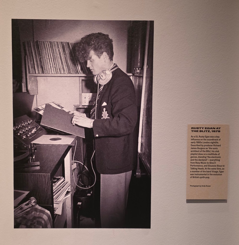 Black and white photograph of a young man, Rusty Egan, in a suit and tie with headphones around his neck, examining a vinyl record in a DJ setup, surrounded by a collection of records.