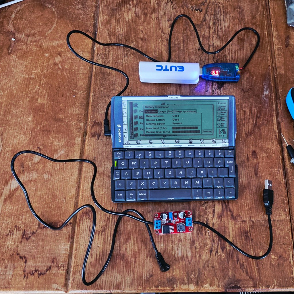 A handheld device with a keyboard displaying battery information on its screen, connected to a power source and a small circuit board on a wooden surface.