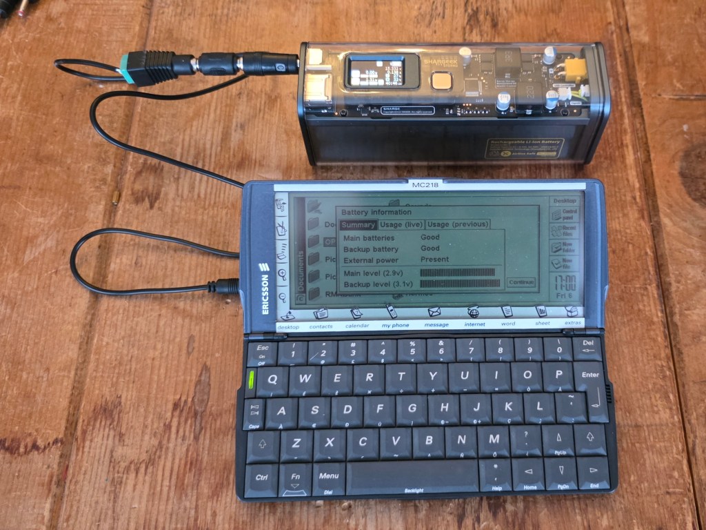 A close-up of an Ericsson MC218 handheld device connected to a battery pack, displaying battery information on the screen. The setup is placed on a wooden surface.