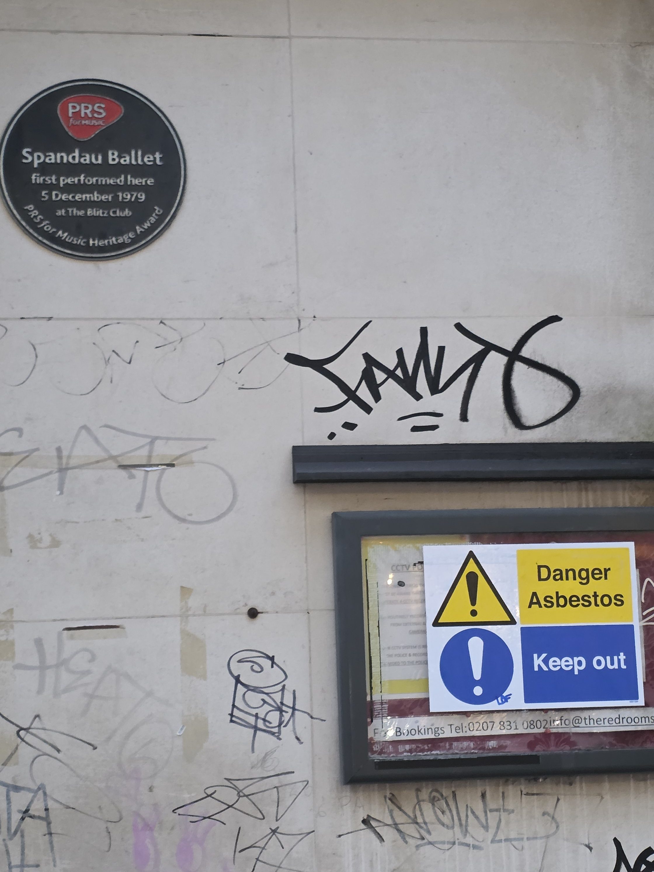 A historic plaque commemorating Spandau Ballet's first performance in 1979, alongside a warning sign about the presence of asbestos, both displayed on a graffiti-covered wall.