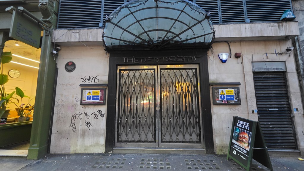 Entrance of a building with a decorative awning labeled 'THE RED ROOMS', featuring metal security doors and graffiti on the walls. Adjacent are signs for a salon and a menu board advertising 'Truffle Raclette'.