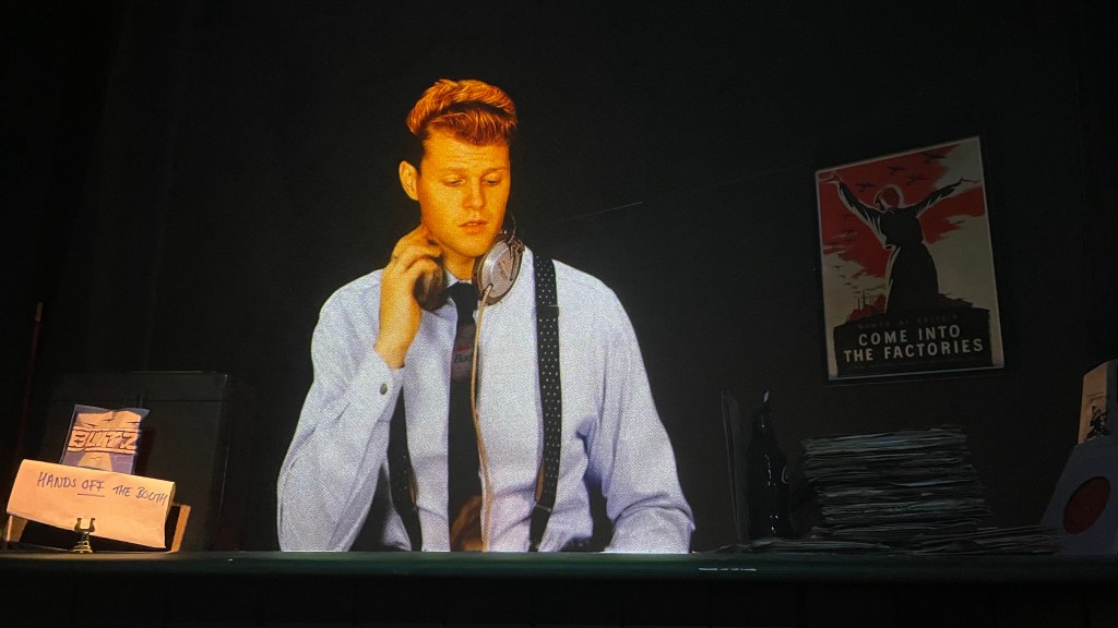 A young man wearing headphones and a button-up shirt is sitting at a desk with a sign that says 'HANDS OFF THE BOOTH'. In the background, there's a propaganda-style poster that reads 'COME INTO THE FACTORIES'.