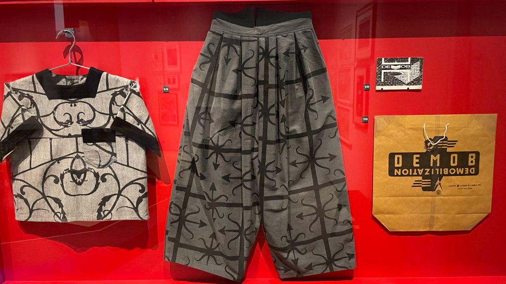 A display featuring a patterned short-sleeve shirt and wide-legged pants hanging on a red background, accompanied by a brown paper bag with a graphic design and the word 'DEMOB' printed on it.
