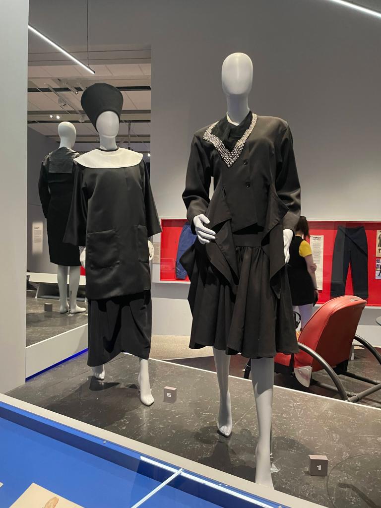 Two black garments displayed on mannequins in a museum setting. The left outfit features a hat-style headpiece with a high collar, while the right outfit has a unique neckline adorned with beads.