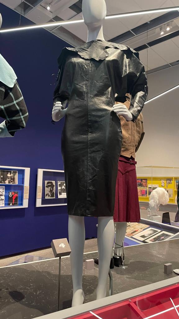 A black leather dress displayed on a mannequin in a museum exhibit, featuring a high collar and puffed sleeves.