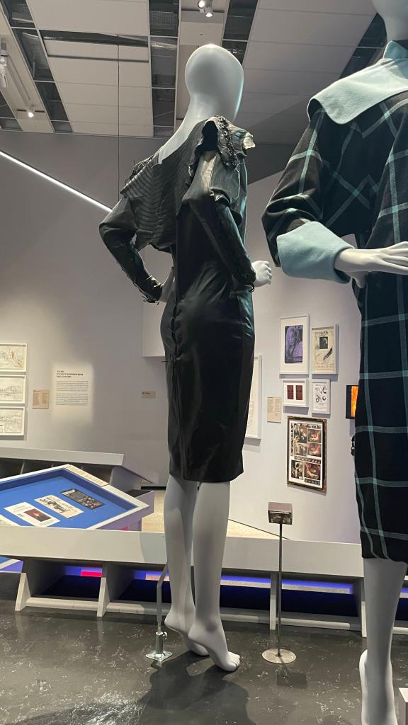 A fashion mannequin displaying a black dress with unique shoulder detailing in an exhibit, surrounded by framed artworks and informational panels.
