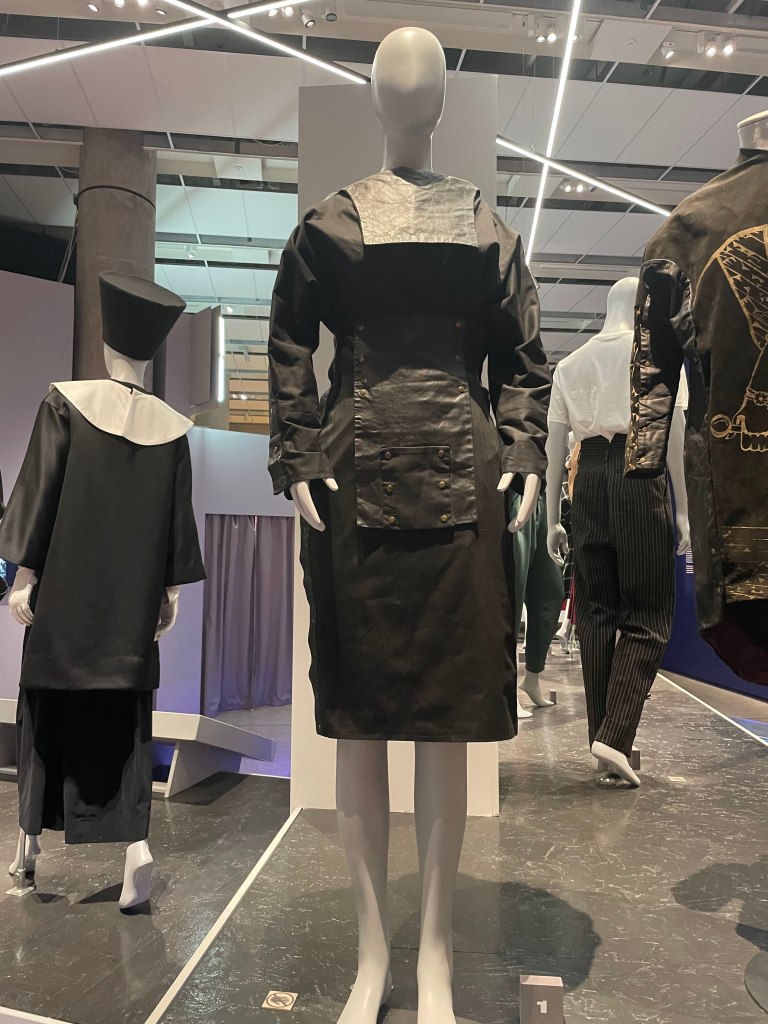 A fashion mannequin displaying a black dress with a contrasting leather panel and button details, surrounded by other garments in a modern exhibition space.