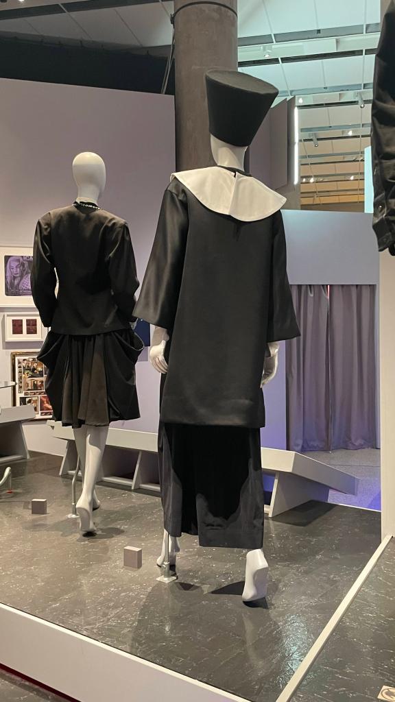 Two mannequins displaying black fashion designs, one featuring a high collar and the other with a structured hat, set in a modern exhibition space.